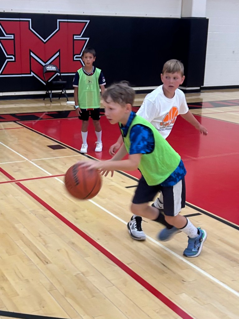 An action shot of a young boy in a lime green scrimmage vest dribbling a basketball down a court during a drill.