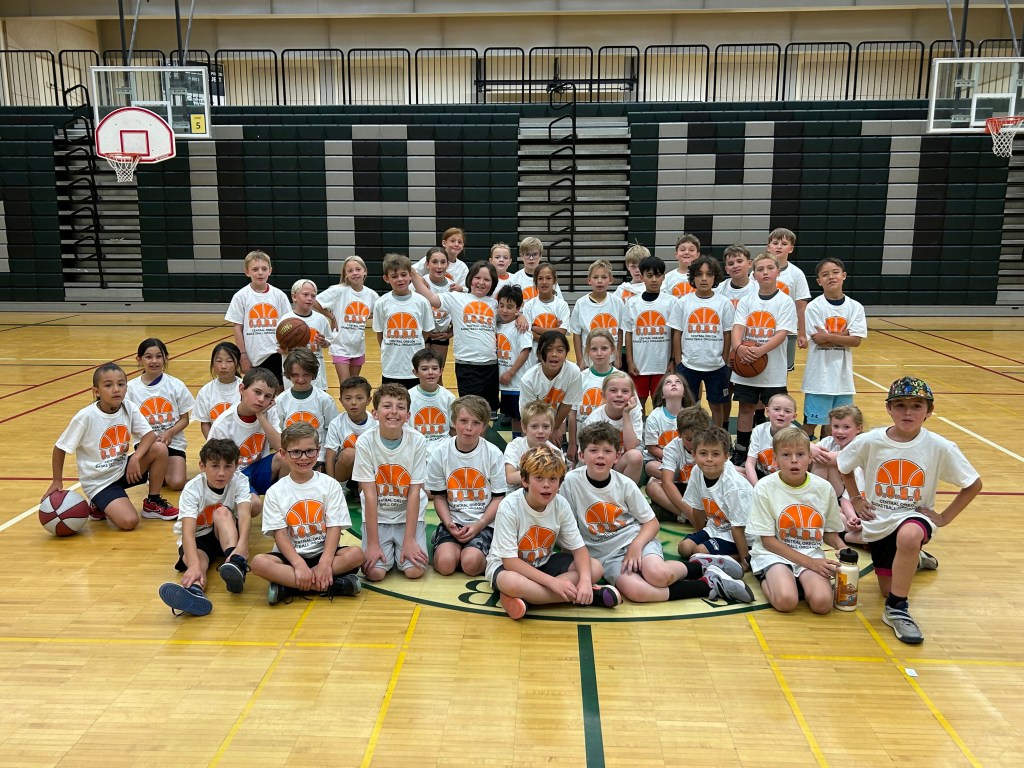 A large group of elementary-aged children wearing matching white "C.O.B.O." basketball t-shirts pose for a group photo on an indoor basketball court.