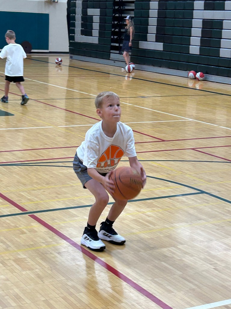 A young boy in a white basketball t-shirt and grey shorts holds a basketball while preparing to take a shot on an indoor court.
